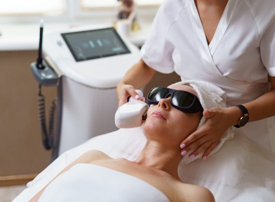 View of doctor cosmetologist doing anti aging procedure in cosmetology office. Satisfied woman in disposable hat lying on couch and relaxing. Working with Apparatus.